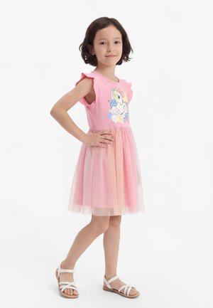 Jeune fille aux cheveux courts et foncés, portant une robe rose avec une licorne et des sandales blanches, debout avec une main sur la hanche.