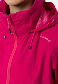 Primo piano di una giacca rosa brillante con colletto alto, chiusura con zip, cappuccio regolabile con lacci e logo Schöffel sul petto.