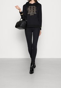 Sudadera negra con un logo dorado, combinada con jeans skinny negros y botas de tobillo negras. El bolso tiene una textura con detalles acolchados.