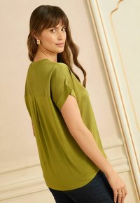 Olívazöld, rövid ujjú blúz laza szabással, kerek nyakkivágással, felhajtott mandzsettákkal és finom ráncolással a hátán.
