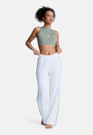 Mujer con pantalones anchos blancos y top corto sin mangas verde posando descalza contra un fondo blanco.