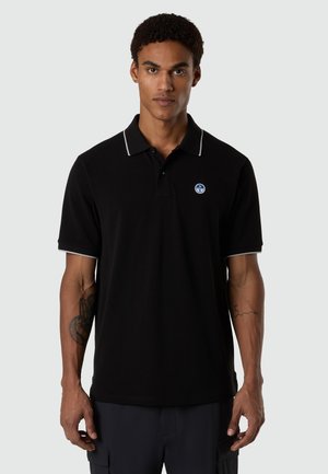 Hombre con camisa tipo polo negra de manga corta con ribete blanco en el cuello y las mangas, de pie con expresión neutral frente a un fondo liso.