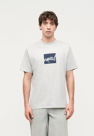 Ung mann med krøllete hår som har på seg en lys grå Rip Curl T-skjorte og lyse grå bukser, står foran en ensfarget lys bakgrunn.