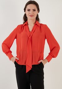 Red blouse in lightweight fabric with a v-neck and a tied bow detail at the collar. Features long sleeves with fitted cuffs and a loose fit.