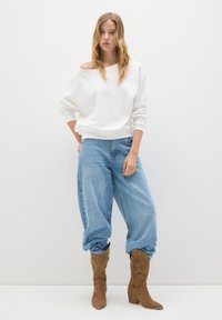 Donna con capelli biondi sciolti, che indossa una felpa bianca scollata, jeans larghi azzurri e stivali in camoscio marroni fino al polpaccio, in piedi contro un muro bianco.
