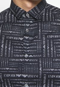 Camisa negra abotonada con texto blanco de Emporio Armani impreso. Cuenta con un cuello estructurado y botones, con una textura textil suave.