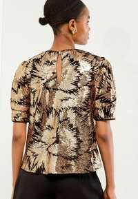 Top a maniche corte con paillettes dorate e bronze luccicanti in un motivo floreale, con un piccolo dettaglio a chiave sulla parte posteriore e una vestibilità rilassata.