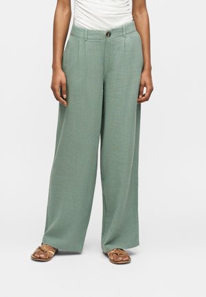 Nő világoszöld, bőszárú nadrágban, gombbal és cipzárral, bézs pántos szandállal és fehér, ráncolt toppal.