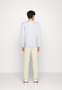 Uomo in piedi con le spalle rivolte alla fotocamera, indossa una camicia bianca a maniche lunghe, pantaloni crema risvoltati alle caviglie, calzini bianchi e sandali marroni.