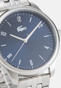 Ceas din metal argintiu cu un cadran texturat de culoare albastru închis, marcaje orare și ace argintii. Prezentă logo-ul crocodil Lacoste la ora 12. Brățară din oțel inoxidabil.