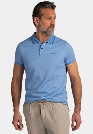 Man met grijze baard, gekleed in een lichtblauw poloshirt en beige broek, kijkt naar zijn rechterkant tegen een effen achtergrond.