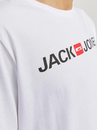 Balta medvilnė marškinėliai su ryškiu juodu "JACK & JONES" užrašu ir raudonu kvadratiniu logotipu. Trumpomis rankovėmis, apvaliu kaklu.