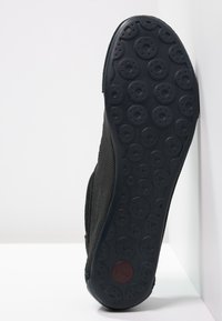 Svart skosula med cirkulära greppmönster och en liten röd cirkulär logotyp nära hälen, vinklad mot en vit hörnbakgrund.