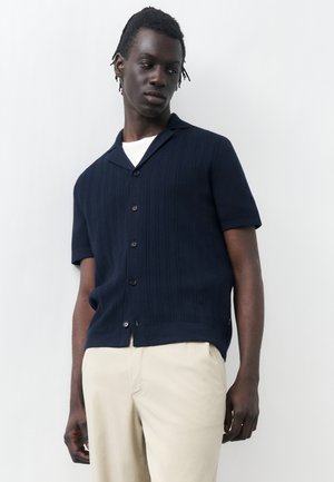 Άνδρας που φοράει σκούρο μπλε κοντομάνικο πουκάμισο με κουμπιά πάνω από λευκό τοπ και μπεζ παντελόνια, στέκεται δίπλα σε λευκό τοίχο και κοιτάζει προς τα κάτω και πλάγια.