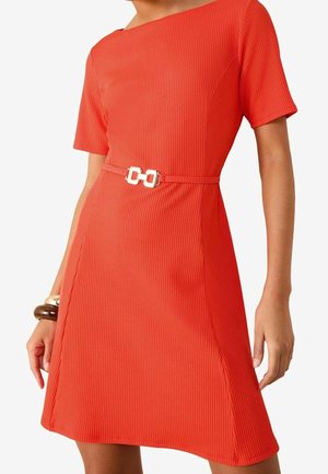 Kobieta ubrana w teksturowaną, krótką, o długości do kolan, czerwoną sukienkę z wąskim paskiem z złotą klamrą oraz bransoletką na lewym nadgarstku.