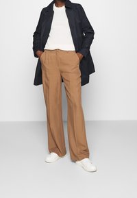 Persona vistiendo pantalones anchos beige, zapatillas blancas, top blanco de canalé y un abrigo trench azul marino con las manos en los bolsillos.