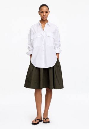 Femme portant une chemise blanche oversize avec manches retroussées, une jupe mi-longue kaki et des tongs noires, debout sur un fond blanc.