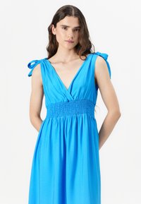 Blå kjole med V-udskæring, smocked talje og bindebånd. Lavet af et let stof, den har en svungne nederdel, der falder elegant.