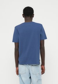 Μπλούζα με κοντά μανίκια σε ναυτικό μπλε, κατασκευασμένη από βαμβάκι, με στρογγυλή λαιμόκοψη και ίσια γραμμή. Η πίσω όψη δείχνει ελάχιστες ραφές και ένα ταμπελάκι στο τελείωμα.