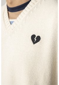 Cremefarbener V-Ausschnitt-Pullover aus Strickstoff mit einem schwarzen zerbrochenen Herzdesign auf der linken Brust. Strukturiertes Material mit geripptem Halsausschnitt.