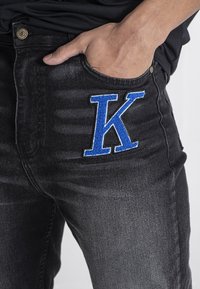 Crne traperice s prednjim džepom koji ima veliki plavi izvezeni detalj slova "K". Materijal ima izbleden izgled.