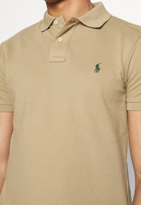 Polo beige in cotone strutturato con una chiusura a tre bottoni e un logo verde sul lato sinistro del petto. Maniche corte con polsini a coste.