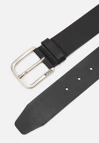 Czarny skórzany pasek z srebrną metalowa klamrą. Wyróżniające cechy to teksturowany pasek oraz tłoczone logo marki w pobliżu klamry.