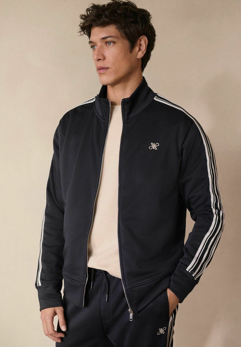Marineblauwe zip-up jas met witte streepaccenten op de mouwen en geborduurd logo op de borst, gecombineerd met bijpassende marineblauwe joggers.