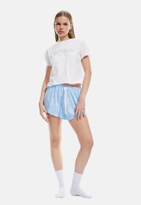 T-shirt bianco corto con grafica blu chiaro, abbinato a pantaloni corti blu con motivi bianchi e vita regolabile con cordino. Calze bianche.