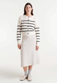 Cárdigan de punto con rayas negras sobre una base blanca, combinado con una falda beige con lunares negros; la modelo lleva zapatos y calcetines claros.