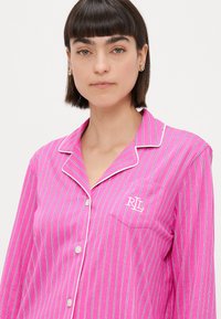 Ροζ ριγέ πιτζάμα με λευκές λεπτομέρειες, κλείσιμο με κουμπιά και τσέπη στο αριστερό στήθος που διαθέτει λογότυπο. Κατασκευασμένη από βαμβακερό ύφασμα με απαλή υφή.