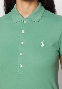 Person i grön pikétröja med vit broderad häst- och ryttarsymbol på vänster bröst, visar krage och knappar.