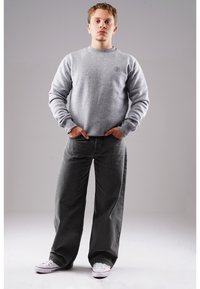 Grauer Pullover mit Rundhalsausschnitt, versehen mit einem dezenten Logo, kombiniert mit locker sitzenden dunkelgrauen Jeans und weißen Sneakers.