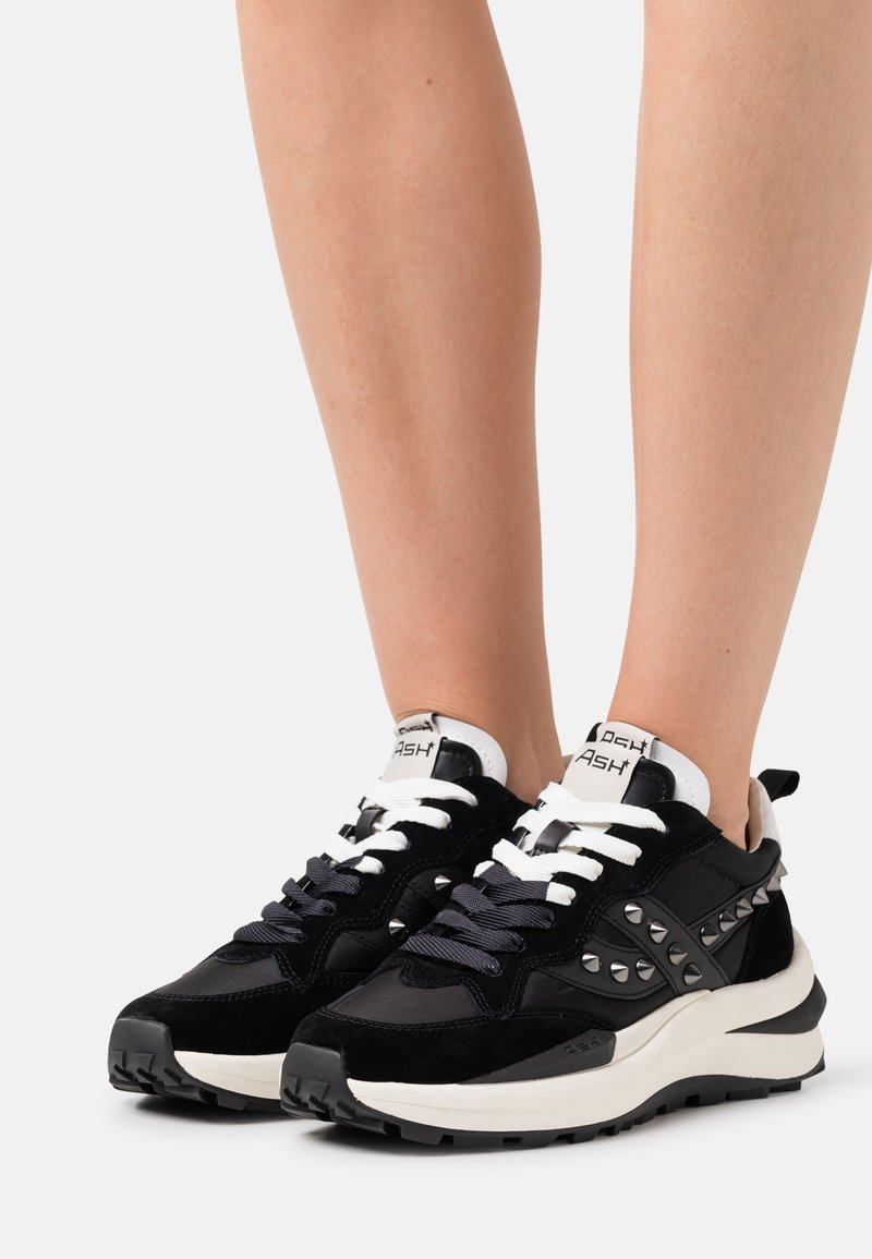 Personne portant des baskets épaisses noires et blanches avec des détails à clous argentés, debout sur un fond clair uni.