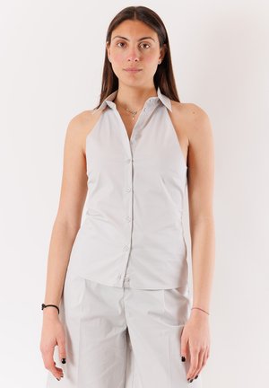 Donna con lunghi capelli scuri che indossa una camicia grigia chiara senza maniche con bottoni e pantaloni grigi chiari abbinati, in piedi davanti a uno sfondo uniforme.