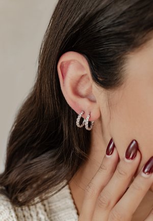 Gros plan sur l'oreille d'une femme portant deux boucles d'oreilles créoles argentées texturées et une main aux ongles manucurés rouge foncé touchant son visage.