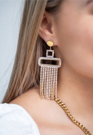 Femme portant une longue boucle d'oreille pendante en or et strass avec des formes géométriques, montrant une partie de la chaîne du collier et un haut blanc texturé.