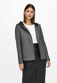 Giacca grigia con texture e cappuccio, con un interno nero liscio. Indossata sopra una camicia bianca e una gonna di pelle nera con uno spacco frontale.