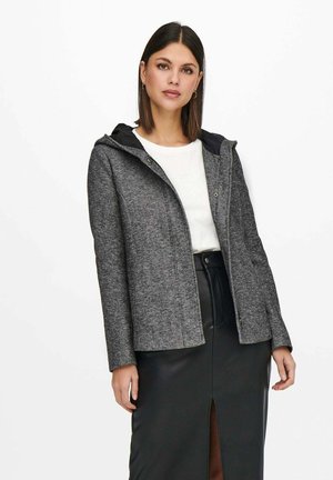 Giacca grigia con texture e cappuccio, con un interno nero liscio. Indossata sopra una camicia bianca e una gonna di pelle nera con uno spacco frontale.