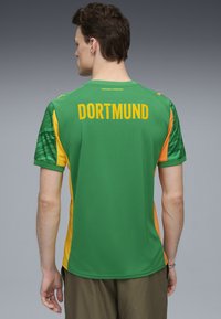 Camisa deportiva verde con detalles amarillos, "DORTMUND" impreso en la parte posterior en letras grandes; cuenta con un patrón texturizado verde en las mangas.