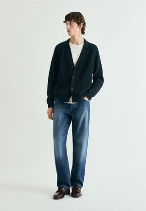 Joven de pie, vistiendo un cárdigan negro sobre una camisa blanca, jeans azules y zapatos de cuero marrón oscuro, con una mano en el bolsillo.