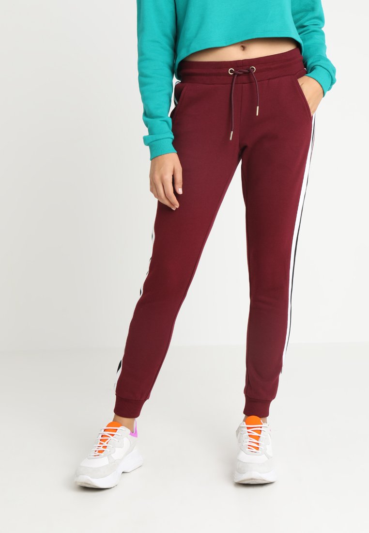Bordeaux joggebukser med elastisk midje og snor, med hvite side-striper. Kombinert med en turkise kortermet genser og hvite joggesko.