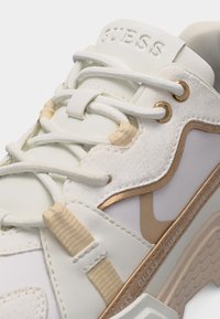 Λευκό και μπεζ αθλητικό παπούτσι Guess με κορδόνια, ανάγλυφο λογότυπο στη γλώσσα και μεταλλικές χρυσές λεπτομέρειες στο πλάι και στα οφθαλμοί.