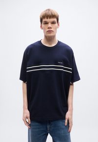 Μπλε ναυτικό βαμβακερό T-shirt με κοντά μανίκια, που διαθέτει μια λευκή οριζόντια ρίγα και το λογότυπο Calvin Klein στο στήθος. Φορεμένο με μπλε τζιν.