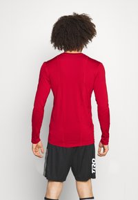 Langärmliges rotes Sporthemd mit glatter Textur, figurbetontem Design und subtilen Nahtdetails, kombiniert mit schwarzen Sportshorts mit weißem Branding.