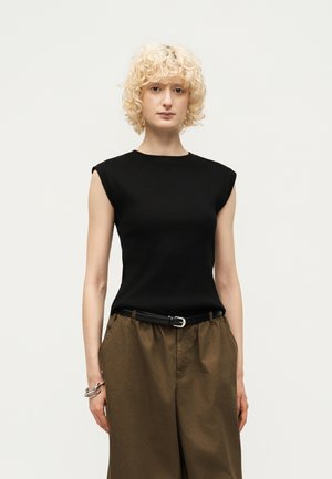 Personne blonde aux cheveux bouclés portant un haut noir sans manches, un pantalon marron taille haute avec une ceinture noire, et des bracelets argentés au poignet gauche.