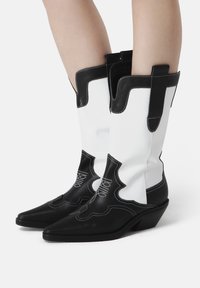 Cizme de cowboy din piele alb-negru, cu vârfuri ascuțite, toc wedge, cusături decorative și mânere de prindere pe țevi.