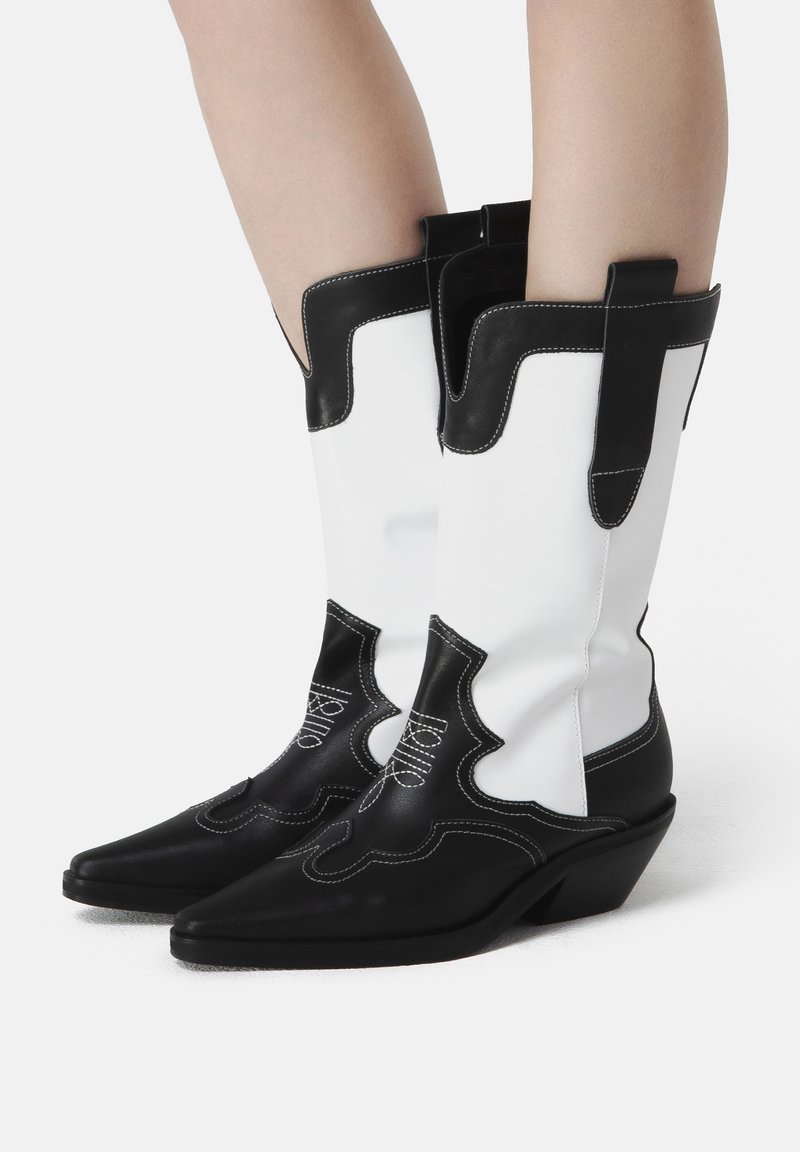 Cizme de cowboy din piele alb-negru, cu vârfuri ascuțite, toc wedge, cusături decorative și mânere de prindere pe țevi.