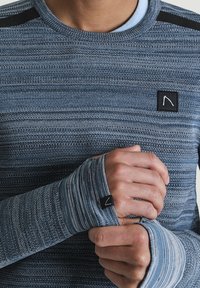 Długi rękaw, niebieska dzianinowa koszula w paski, z czarnymi akcentami na ramionach i małą czarnąłatką z logo na klatce piersiowej.