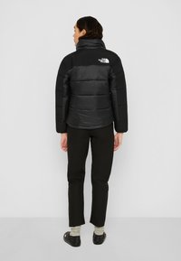 Svart pufferjakke med høy krage og horisontalt stripe-design, med The North Face-logoen på baksiden. Brukes med svarte bukser.